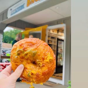 May include: A hand holds a freshly baked bagel with melted cheese and green peppers. The bagel is golden brown with a slightly crispy exterior. The sign above reads "Lakeshore Bagel".