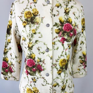 May include: Cream-colored floral jacket with a round neckline and three-quarter sleeves. The jacket features a pattern of yellow and pink roses with green foliage. It has decorative silver buttons down the front.