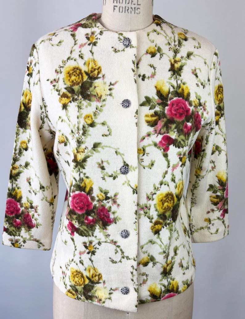 May include: Cream-colored floral jacket with a round neckline and three-quarter sleeves. The jacket features a pattern of yellow and pink roses with green foliage. It has decorative silver buttons down the front.