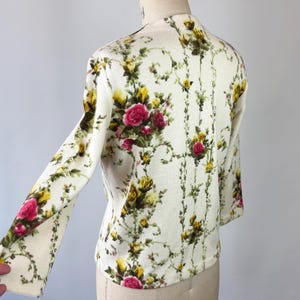 May include: Cream-colored floral print cardigan with long sleeves. The pattern features pink and yellow roses with green foliage on a cream background. The cardigan is displayed on a mannequin.