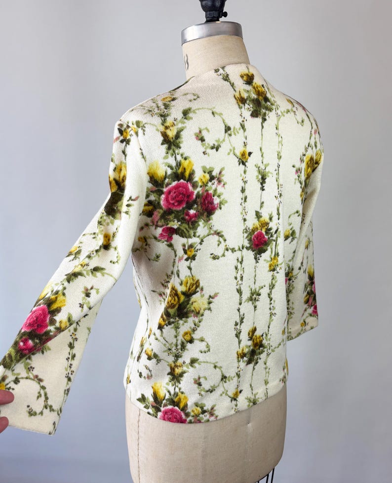 May include: Cream-colored floral print cardigan with long sleeves. The pattern features pink and yellow roses with green foliage on a cream background. The cardigan is displayed on a mannequin.
