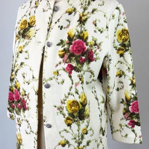 May include: Cream-colored floral jacket with a button-up front. The jacket features a pattern of yellow and pink roses with green foliage. The sleeves are three-quarter length, and the jacket is displayed on a mannequin.