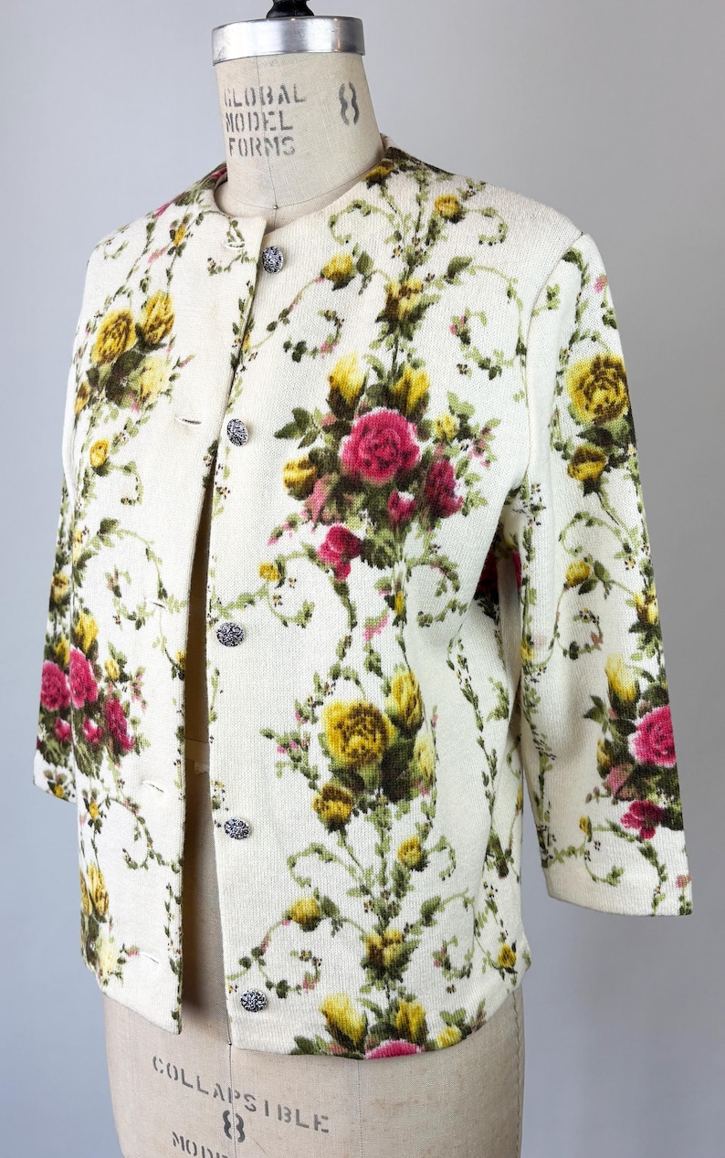 May include: Cream-colored floral jacket with a button-up front. The jacket features a pattern of yellow and pink roses with green foliage. The sleeves are three-quarter length, and the jacket is displayed on a mannequin.