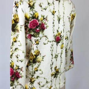 May include: Cream-colored floral cardigan with a vibrant pattern of red, yellow, and green roses and foliage. The long-sleeved sweater is displayed on a mannequin, showcasing the intricate design and texture of the knitwear.