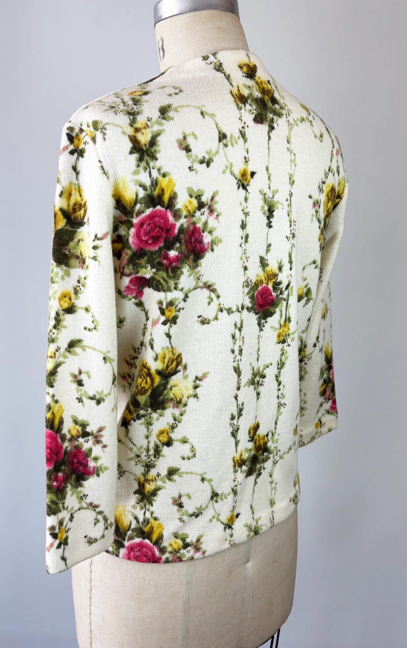 May include: Cream-colored floral cardigan with a vibrant pattern of red, yellow, and green roses and foliage. The long-sleeved sweater is displayed on a mannequin, showcasing the intricate design and texture of the knitwear.