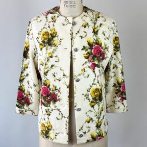 May include: Cream-colored jacket with a floral pattern of yellow and pink roses and green vines. The jacket has a round collar, three-quarter sleeves, and decorative buttons. The garment is displayed on a mannequin.