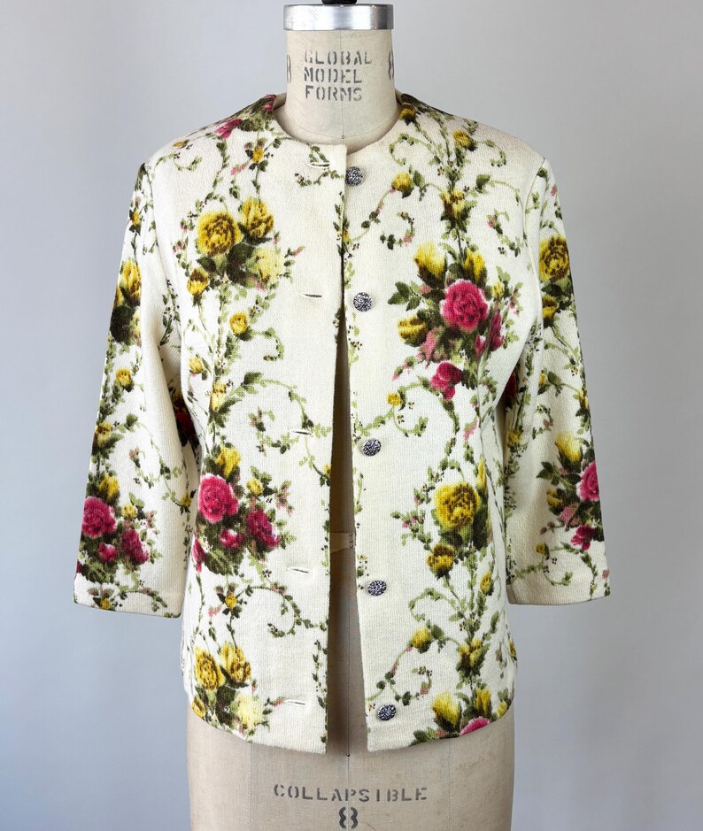 May include: Cream-colored jacket with a floral pattern of yellow and pink roses and green vines. The jacket has a round collar, three-quarter sleeves, and decorative buttons. The garment is displayed on a mannequin.