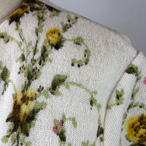 May include: Cream-colored sweater with a floral pattern. The design features yellow and green flowers with green vines and small pink accents. The sweater has a crew neck and a knitted texture.