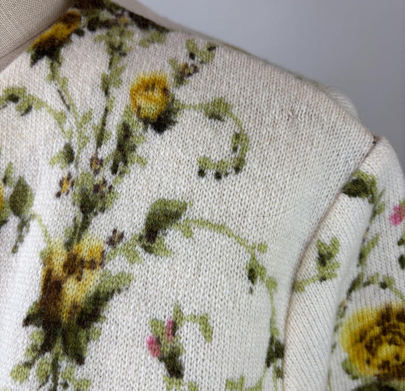 May include: Cream-colored sweater with a floral pattern. The design features yellow and green flowers with green vines and small pink accents. The sweater has a crew neck and a knitted texture.