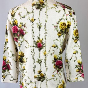 May include: Cream-colored cardigan with a floral pattern featuring yellow and red roses, green leaves, and vines. The three-quarter sleeve sweater is displayed on a mannequin. The floral design covers the entire garment.