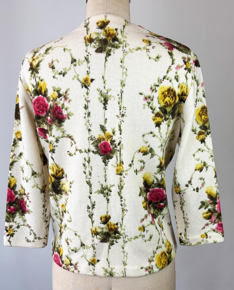 May include: Cream-colored cardigan with a floral pattern featuring yellow and red roses, green leaves, and vines. The three-quarter sleeve sweater is displayed on a mannequin. The floral design covers the entire garment.