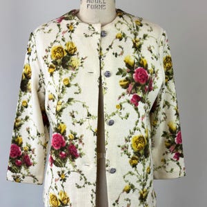 May include: Cream-colored jacket with a floral pattern of yellow and red roses and green leaves. The jacket has three-quarter sleeves and is buttoned with silver-colored buttons. The jacket is displayed on a mannequin.