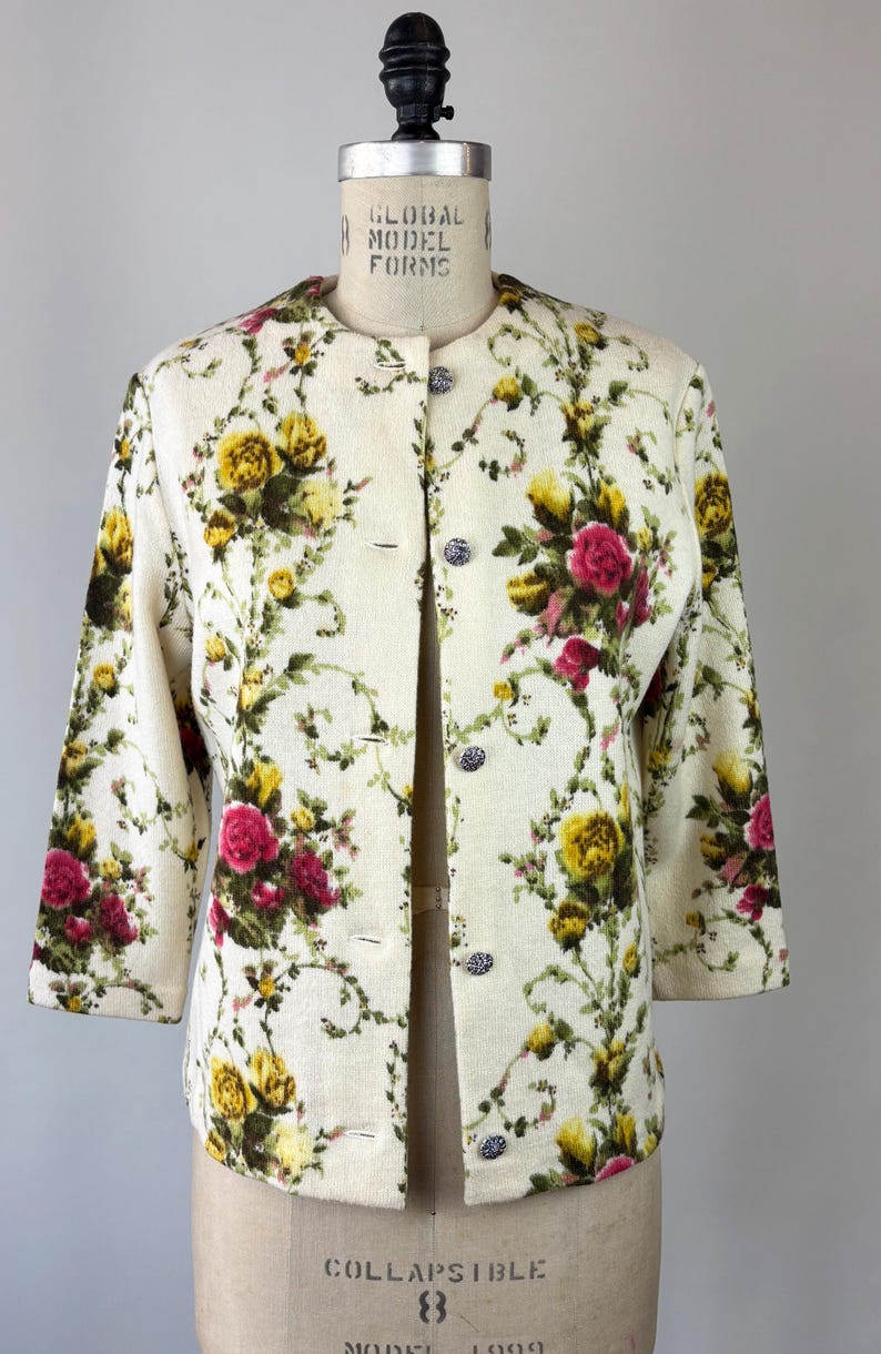 May include: Cream-colored jacket with a floral pattern of yellow and red roses and green leaves. The jacket has three-quarter sleeves and is buttoned with silver-colored buttons. The jacket is displayed on a mannequin.