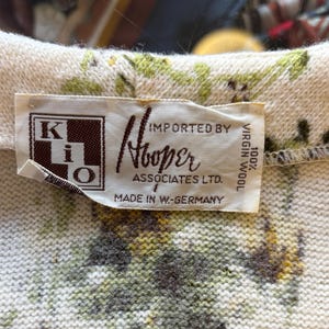 May include: Close-up of a vintage sweater collar with a floral pattern in green, yellow, and brown on a cream background. The label reads "Hooper Associates Ltd." and "100% Virgin Wool."