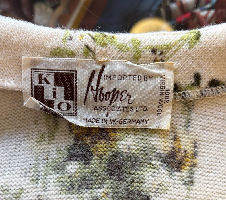 May include: Close-up of a vintage sweater collar with a floral pattern in green, yellow, and brown on a cream background. The label reads "Hooper Associates Ltd." and "100% Virgin Wool."