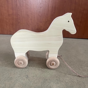 May include: A wooden pull toy shaped like a horse with a light brown finish. The toy has four wheels and a rope attached to the horse's head.
