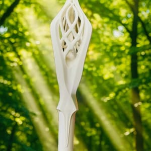 May include: A white, intricately carved staff with a stylized floral or leaf-like design at the top. The staff has a long, cylindrical body and is set against a blurred green forest background.