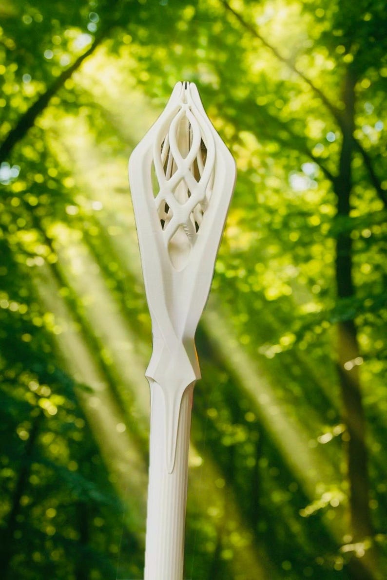 May include: A white, intricately carved staff with a stylized floral or leaf-like design at the top. The staff has a long, cylindrical body and is set against a blurred green forest background.