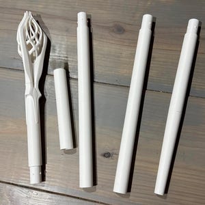 May include: A collection of white 3D-printed components, including a decorative, ornate top piece and several cylindrical rods of varying lengths. The pieces are arranged on a gray wooden surface.