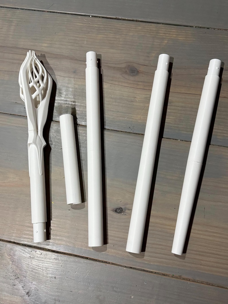 May include: A collection of white 3D-printed components, including a decorative, ornate top piece and several cylindrical rods of varying lengths. The pieces are arranged on a gray wooden surface.
