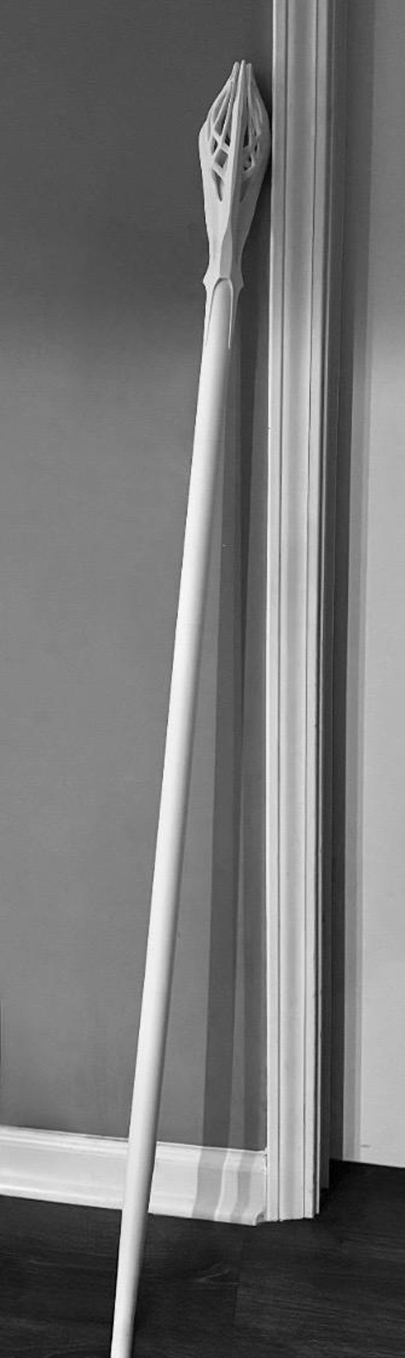 May include: A tall, white staff with a detailed, geometric top design. The staff leans against a wall and door frame, showcasing its length and intricate craftsmanship. The staff appears to be made of wood or a similar material.