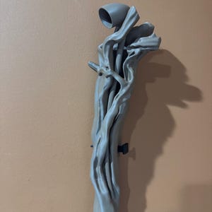 May include: A gray, abstract sculpture resembling a tree branch with a cup-like structure at the top. The sculpture has a textured surface and is mounted on a wall. The overall design is organic and artistic.