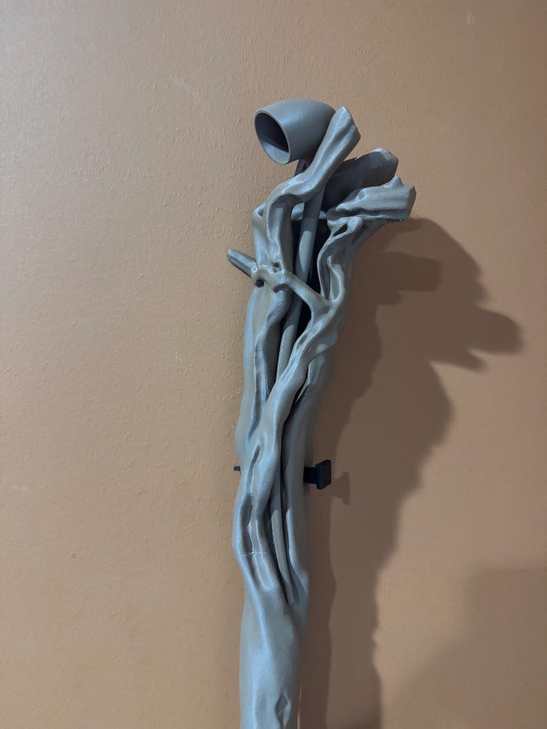 May include: A gray, abstract sculpture resembling a tree branch with a cup-like structure at the top. The sculpture has a textured surface and is mounted on a wall. The overall design is organic and artistic.