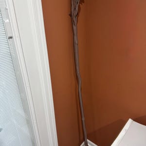 May include: A brown wooden staff with a detailed, textured top and a smooth, slender body. The staff is leaning against a brown wall, showcasing its intricate design and craftsmanship. The staff is approximately 60 inches tall.