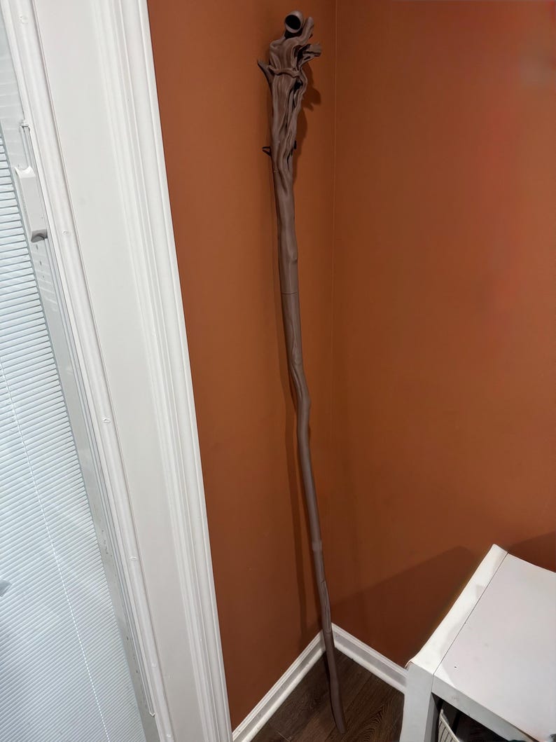May include: A brown wooden staff with a detailed, textured top and a smooth, slender body. The staff is leaning against a brown wall, showcasing its intricate design and craftsmanship. The staff is approximately 60 inches tall.