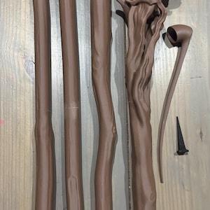 May include: Brown wooden staff components, including four cylindrical pieces, a detailed top section, a curved piece, and a small black spike. The pieces are arranged on a wooden surface.
