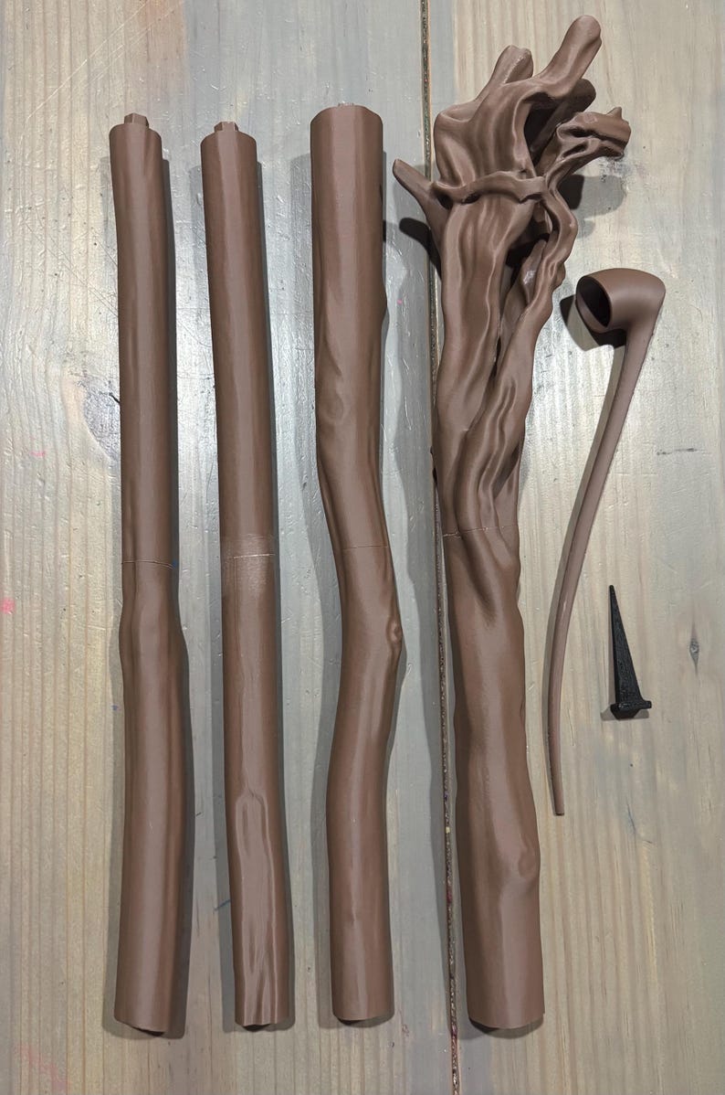 May include: Brown wooden staff components, including four cylindrical pieces, a detailed top section, a curved piece, and a small black spike. The pieces are arranged on a wooden surface.