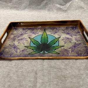 May include: A wooden serving tray with a clear glass insert featuring a green marijuana leaf design on a blue background. The design is surrounded by purple marijuana leaf outlines.