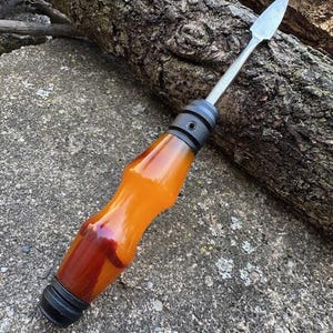 May include: A handcrafted tool with a polished, amber-colored handle and a metal blade. The handle transitions to a deep red hue near the base, with black accents. The tool rests on a gray stone surface, with a log in the background.