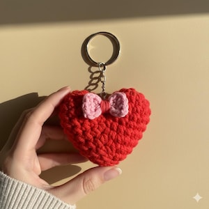 Puede incluir: Un llavero de corazón rojo tejido a mano con un lazo rosa. El dije en forma de corazón está unido a un llavero plateado. Un accesorio encantador.
