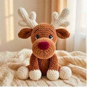 May include: A handmade brown crochet reindeer with white antlers, ears, and foot pads. The reindeer has a red nose and black eyes. The plush toy is sitting on a cream-coloured knitted blanket.