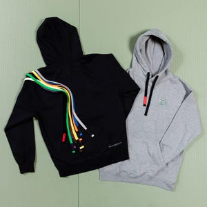 May include: Two hooded sweatshirts, one black and one gray. The black sweatshirt has a rainbow of colorful strings hanging from the back. The gray sweatshirt has a small logo on the front. Both sweatshirts have drawstrings.