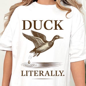 Pode incluir: Camiseta branca com um gráfico marrom de um pato em voo, ondulações de água e o texto "DUCK LITERALLY". A camiseta é uma Gildan 5000 White.