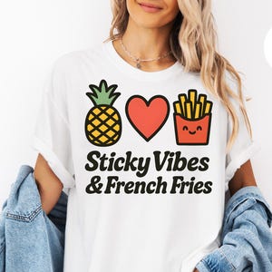 Puede incluir: Camiseta blanca con un diseño gráfico que presenta una piña, un corazón y una caja de patatas fritas. El texto "Sticky Vibes & French Fries" está impreso debajo. La camisa se usa con una chaqueta vaquera.