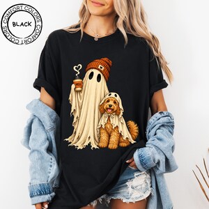May include: A black t-shirt featuring a graphic of a ghost wearing an orange beanie and holding a coffee cup with the text MEH, standing next to a golden-brown dog wearing a ghost costume. A circular logo in the corner reads COMFORT COLORS BLACK.