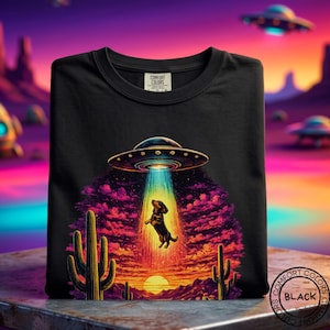 Könnte beinhalten: Ein schwarzes T-Shirt mit einer Grafik eines Dackels, der von einem UFO in einer Wüstenlandschaft mit Kakteen, einem Sonnenuntergang und violetten Wolken entführt wird. Das T-Shirt hat ein Comfort Colors-Etikett am Innenkragen und ein schwarzes Etikett.