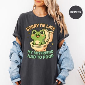May include: Dark gray t-shirt with a humorous graphic. The design features a cartoon frog sitting on a toilet, with the text "SORRY I'M LATE" above and "MY BOYFRIEND HAD TO POOP" below. The frog is green, and the toilet is beige.