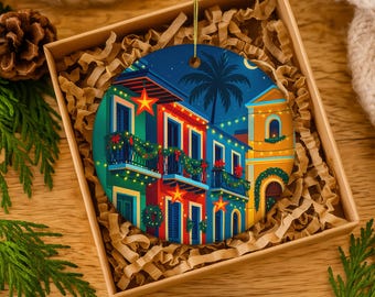 May include: A round Christmas ornament featuring a colorful illustration of buildings decorated with lights and wreaths. The buildings are painted in shades of blue, red, and yellow, with a palm tree and a night sky background. The ornament is in a gift box.