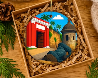 May include: A round Christmas ornament featuring a colorful illustration of a red building with an open door, palm trees, and a stone tower. The ornament is displayed in a brown gift box with shredded paper.