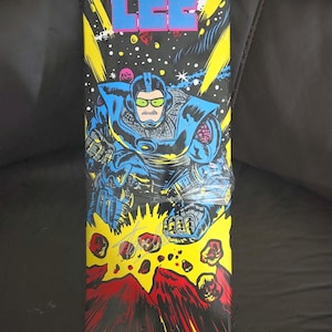May include: A black and yellow skateboard deck with a cartoon image of Stan Lee in a blue spacesuit. The deck features a red and yellow volcano with a black background. The text "Stan Lee" is printed in blue and pink on the top of the deck. The text "Stan Lee Collectibles.com" is printed in white on the bottom of the deck. The number "175" is printed in white on the bottom of the deck.