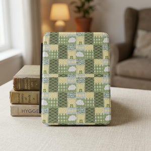 Op de afbeelding: Een rechthoekige e-reader hoes met een patchworkpatroon van groene, gele en witte vierkanten. Het ontwerp bevat herhalende illustraties van kleine groene kikkers, witte wolken, geblokte rasters en ruitvormige lijnen over het oppervlak.