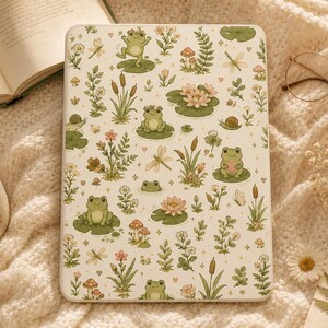May include: A rectangular e-reader cover featuring a repeating pattern of green frogs, pink water lilies, mushrooms, dragonflies, snails, and various wildflowers on a cream background. The design includes small heart and star accents.