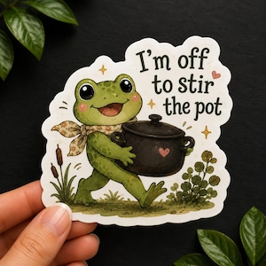 Può includere: Un adesivo fustellato raffigurante una rana verde con una sciarpa a fiori che trasporta una pentola nera. Il testo recita, I'm off to stir the pot. L'illustrazione include piccoli cuori rosa, stelle gialle, tife e fogliame verde.
