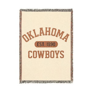 May include: Cream-coloured woven throw blanket with brown text that reads "OKLAHOMA COWBOYS EST. 1890". The blanket has a fringed edge in a matching brown colour. The blanket is rectangular.