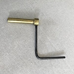 Puede incluir: Una herramienta en forma de L de lat&oacute;n y metal negro con un mango redondo. La herramienta se utiliza para apretar o aflojar tornillos.