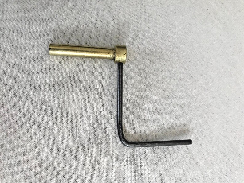 Puede incluir: Una herramienta en forma de L de lat&oacute;n y metal negro con un mango redondo. La herramienta se utiliza para apretar o aflojar tornillos.